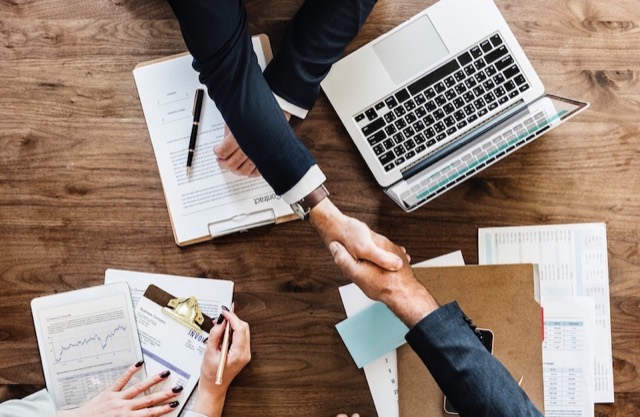 Twee mensen geven elkaar een hand boven een tafel met een laptop en papieren
